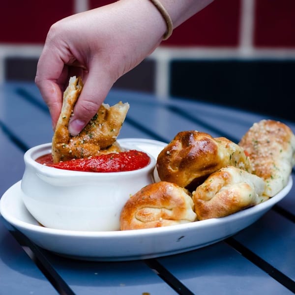 Hand dipping a garlic knot into a white bowl of marinara sauce on a dark blue table.