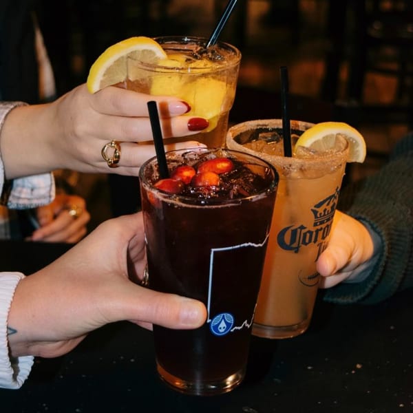 Three hands holding up different cocktails, including a dark drink with cranberries and two lemon-garnished drinks.
