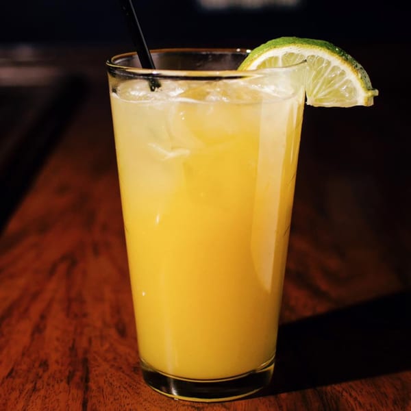 A tall glass of yellow cocktail with ice, a black straw, and a lime wedge on a wooden table.