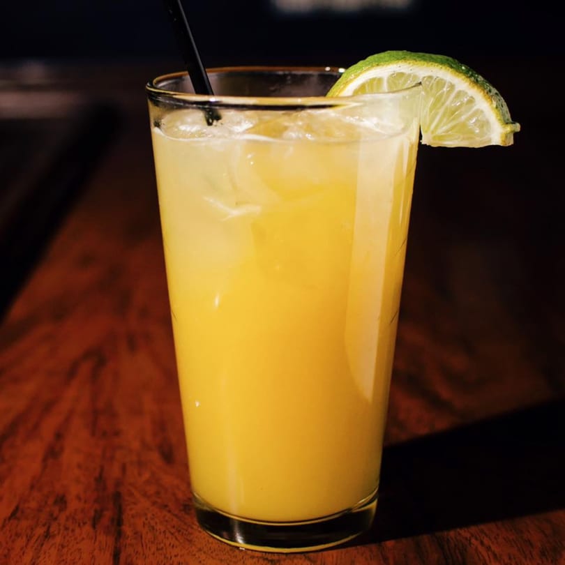 A tall glass of yellow cocktail with ice, a black straw, and a lime wedge on a wooden table.