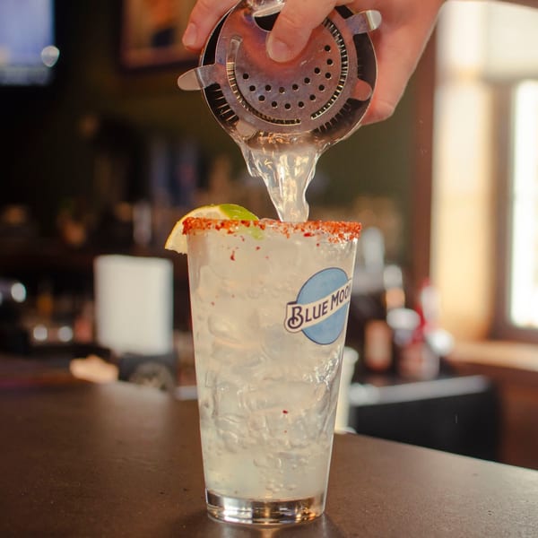A bartender pours a drink from a shaker into a Blue Moon glass with a chili-lime rim.