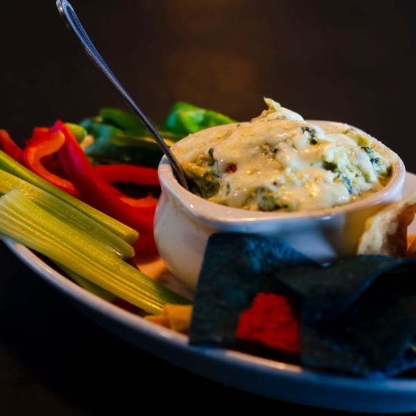 Creamy spinach dip with melted cheese, served with fresh celery sticks, red bell peppers, and tortilla chips.